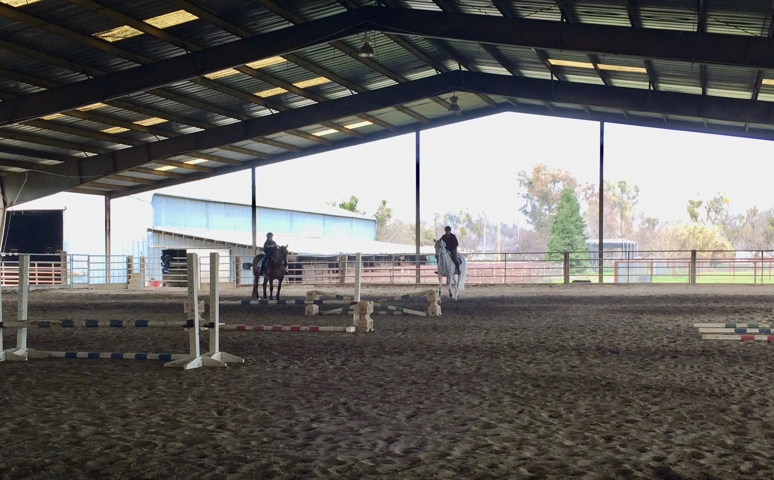 Two riders in the covered arena with jump standards set up